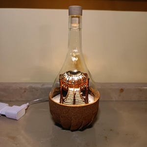 Può includere: Una bottiglia di vetro decorativa con tappo in sughero, che ospita una struttura in miniatura illuminata. La bottiglia ha una base in sughero marrone e un corpo in vetro trasparente. L'interno presenta un modello architettonico dettagliato e illuminato.