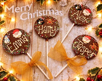 Könnte beinhalten: Drei runde Schokoladenlutscher an weißen Stielen, verziert mit den in Zuckerguss geschriebenen Namen Emma, Harry, Alex und Jasper. Die Lutscher zeigen essbare Zuckerfiguren von Weihnachtsmännern, Schneemännern, Rentieren und Pinguinen mit Streuseln.