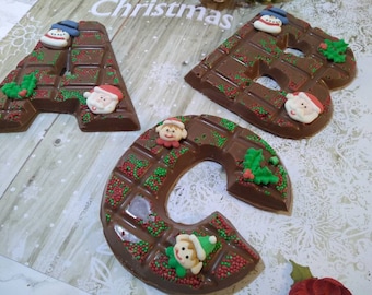 May include: Chocolate letters A, B, and C, decorated for Christmas. Each letter is made of dark chocolate and adorned with festive sprinkles, holly, and Santa Claus and snowman figures. The letters are on a light wood-grain surface.