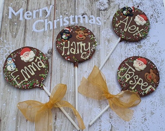 May include: Chocolate lollipops with Christmas-themed decorations and personalized names. Each lollipop features a round chocolate base with festive designs like Santa Claus, reindeer, and a snowman, along with the names "Emma", "Harry", "Alex", and "Jasper". Gold ribbons add a decorative touch.