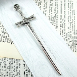May include: A silver sword bookmark with a cross design on the hilt and blade. The bookmark is on a white surface with a book in the background.