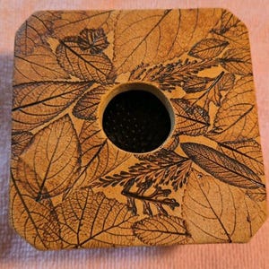 May include: A square, tan-colored ceramic flower frog featuring a detailed, dark-etched pattern of various overlapping leaves across its top surface. The object has a circular opening in the center that reveals a dark, spiked metal pin holder.