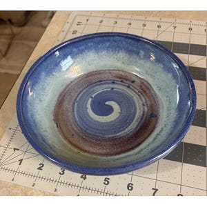 May include: A handmade ceramic bowl with a spiral design. The bowl features a dark blue rim, transitioning to light blue and white, with a central spiral in shades of purple and brown. The bowl is approximately 8 inches in diameter.
