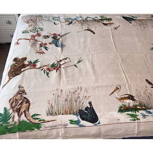 May include: A beige fabric tablecloth featuring a printed pattern of Australian wildlife, including a kangaroo, a koala, a black swan, and a pelican. The text "Australian Wildlife" is printed in the lower section of the fabric.