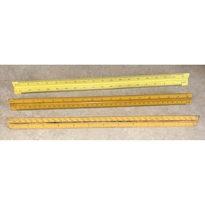 May include: Three triangular architectural scales arranged horizontally. The top scale is bright yellow with black markings. The middle scale is a darker, aged wood tone with black markings. The bottom scale is a light tan wood with black markings.