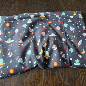 May include: A dark grey pillowcase featuring a colourful space-themed pattern. The design includes rockets, planets, aliens, stars, and moons in red, yellow, blue, and green. The pillowcase is made of soft fabric.
