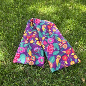 May include: A rectangular fabric heating pad featuring a vibrant floral pattern on a deep purple background. The design includes stylised flowers and leaves in shades of bright pink, orange, yellow, and teal, arranged in a repeating botanical print.