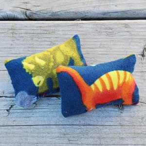 May include: Two blue fabric cushions with dinosaur designs. One cushion features a yellow dinosaur and the other features an orange dinosaur.