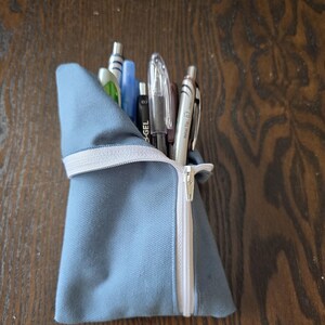 May include: A light blue fabric pencil case with a white zipper and a white elastic band. The case is filled with various pens and pencils. The case is sitting on a dark wooden surface.