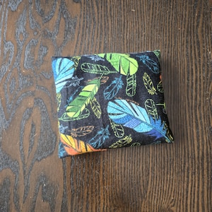 Peut inclure: Petit coussin carré avec un motif de plumes colorées sur fond noir. Les plumes sont bleues, vertes, jaunes et oranges. Le coussin est posé sur une surface en bois.