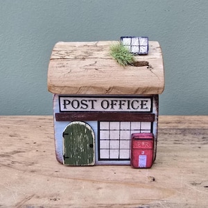 May include: A miniature wooden post office model with a light brown roof, a green door, and a red mailbox. The sign above the window reads "POST OFFICE" in black letters. The building is painted in shades of blue and white.
