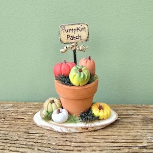 May include: A miniature pumpkin patch decoration featuring a terracotta pot filled with colorful pumpkins and greenery. A sign that reads "Pumpkin Patch" is displayed above the pumpkins. The arrangement sits on a wooden slice base.