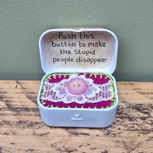 May include: A small, pale blue tin with a hinged lid, inscribed with the words "Push this button to make the stupid people disappear." Inside, a pink button sits on magenta fabric with white lace trim.