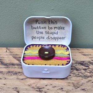 May include: A white box with a hinged lid, featuring the text "Push this button to make the stupid people disappear." Inside, a brown button is set on yellow felt, with green and pink ribbons. The box rests on wood.