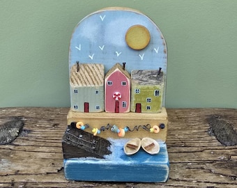 May include: Wooden seaside diorama featuring three colorful houses with textured roofs, a blue sky with a yellow sun, and white birds. Below, a blue sea with a boat and two small boats. Decorative nautical theme.