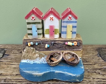May include: A decorative wooden beach scene featuring three miniature beach huts with red roofs, striped walls, and colorful doors. The scene includes a blue ocean, a small wooden island, and two walnut shell boats. Decorative accents include small beads and wire.