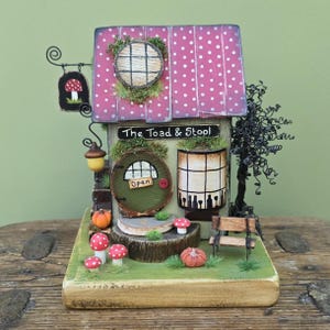 May include: A miniature wooden house painted green with a red and white polka dot roof. The house has a sign that reads "The Toad & Stool" and a door with a sign that says "Open". There are mushrooms, pumpkins, and a small bench in front of the house.