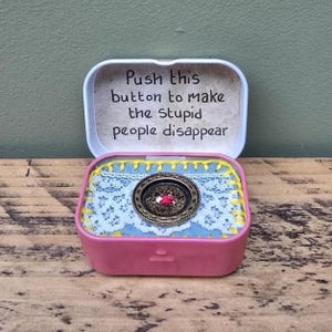 May include: A pink metal box with a hinged lid, containing a blue fabric base with a lace overlay and a decorative button. The lid has a handwritten note that reads, "Push this button to make the stupid people disappear."