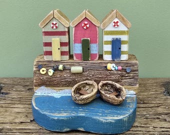 May include: A wooden beach-themed decorative piece featuring three miniature beach huts with red, pink, and green stripes. The huts sit on a wooden base with a blue wave design, and two walnut shell boats.