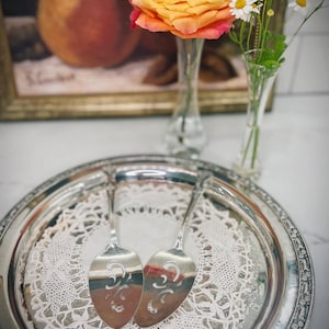 May include: A silver serving tray with two matching cake servers, resting on a white lace doily. The tray is decorated with an ornate border. A vase with a yellow and pink rose and a vase with daisies are in the background.