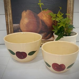 May include: Two vintage ceramic mixing bowls with a cream colored glaze and a red apple design. The bowls are nested together.