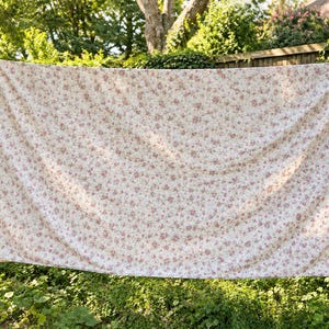 May include: A large rectangular fabric sheet featuring an all-over floral pattern with small pink and red blossoms on a cream-colored background. The textile is draped horizontally, displaying soft folds and a consistent repeating botanical print.