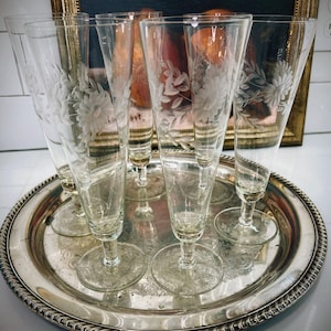 May include: Six clear glass champagne flutes with etched floral designs sit on a silver serving tray.