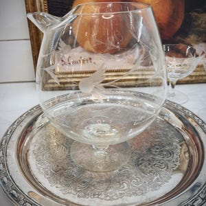 May include: A clear glass decanter with an etched design of birds and leaves sits on a silver tray with an ornate design. The decanter has a spout and a round base.
