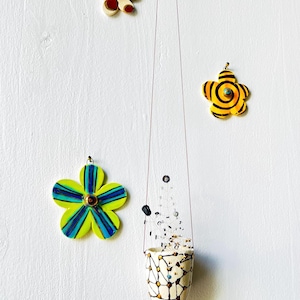 May include: A collection of handmade floral ornaments and a hanging ceramic planter against a white wall. The ornaments feature bright colors and unique designs, including yellow, blue, and brown. The planter is white with a black and gold pattern.