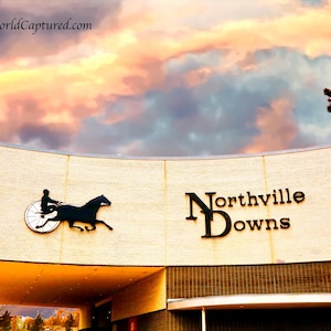 May include: Exterior shot of Northville Downs, a horse racing venue, under a vibrant sunset. The building features a curved facade with the venue's name in large, black letters and a silhouette of a horse and driver.
