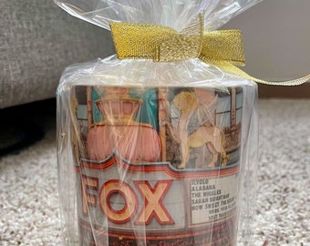 May include: A white ceramic mug with a colorful image of a vintage movie theater marquee with the word "FOX" in red and yellow letters. The mug is wrapped in clear plastic with a gold ribbon.