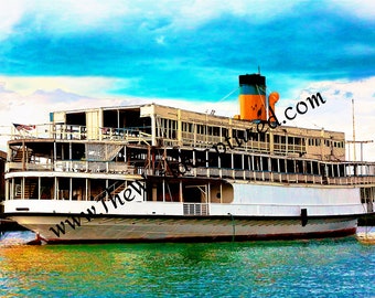 May include: A large, white, wooden ferry boat with a yellow and blue smokestack. The boat is docked at a pier and the water is a turquoise color. The sky is blue with white clouds.