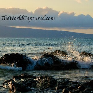 May include: A scenic ocean view at sunset. Waves crash against dark rocks in the foreground, with a mountain range and cloudy sky in the background. The website address www.TheWorldCaptured.com is visible.
