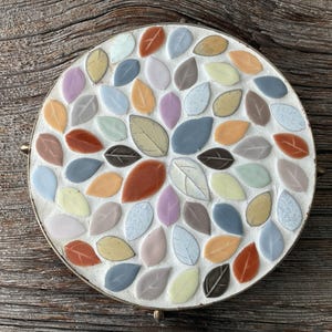 May include: A round trivet with a mosaic leaf design. The leaves are in various colors, including orange, blue, yellow, and brown. The trivet has a white border and gold-colored handles. The trivet is on a wooden surface.