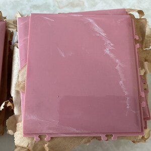 May include: A stack of rectangular, dusty rose-colored plastic tiles with a smooth, glossy finish and subtle white marbled veining. Each tile features small, notched cutouts along the edges, designed for interlocking assembly or mounting.