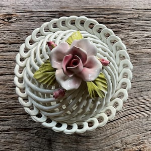 May include: A white ceramic basket with a woven design, adorned with a pink and red rose and green leaves. The decorative piece has a circular shape and is set against a wooden background.