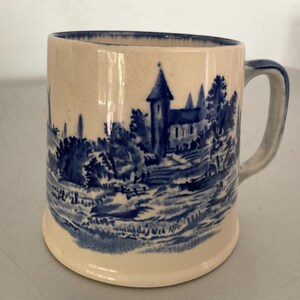 May include: A white ceramic mug with a blue and white landscape design. The design features a church, trees, and a river. The mug has a handle.