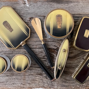 May include: A collection of vintage vanity items, including a hand mirror, compacts, and a brush. The pieces feature a gold and black color scheme with Art Deco designs. The items are arranged on a wooden surface.