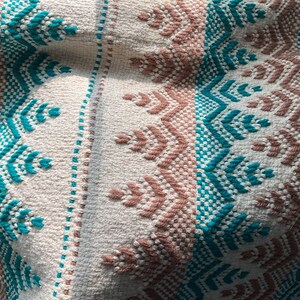 May include: Close-up of a woven textile with a repeating pattern. The design features alternating bands of white, teal, and brown geometric shapes. The texture appears soft and the colors are muted, creating a cozy feel.