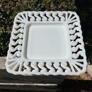 May include: A square, white, decorative dish with a detailed, openwork design around the edges. The dish has a smooth, flat center and a scalloped, scroll-like pattern on the border. The dish is made of a solid, opaque material.