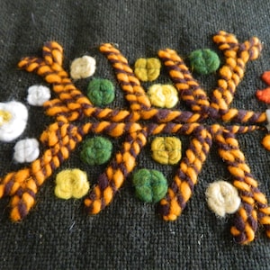 May include: Close-up of a dark green fabric with colorful embroidery. The embroidery features a central design of twisted orange and brown threads, surrounded by small green, yellow, and white floral accents. The overall texture is dense and detailed.