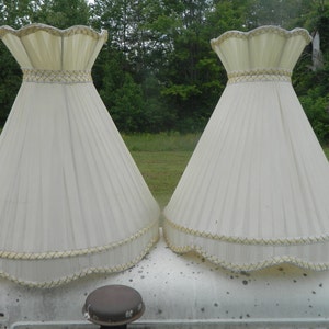 May include: Two off-white lampshades with a pleated design. Each lampshade has a scalloped top and bottom edge, trimmed with gold and cream-colored accents. The lampshades are cone-shaped and sit on a white surface.