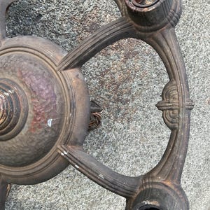 May include: An antique, bronze-colored chandelier frame with three bulb sockets. The frame has an ornate, circular design with decorative flourishes and a textured surface. The metal shows signs of age and wear.