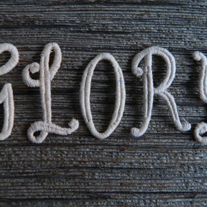 May include: The word "GLORY" is embroidered in white thread on a rustic wooden surface.