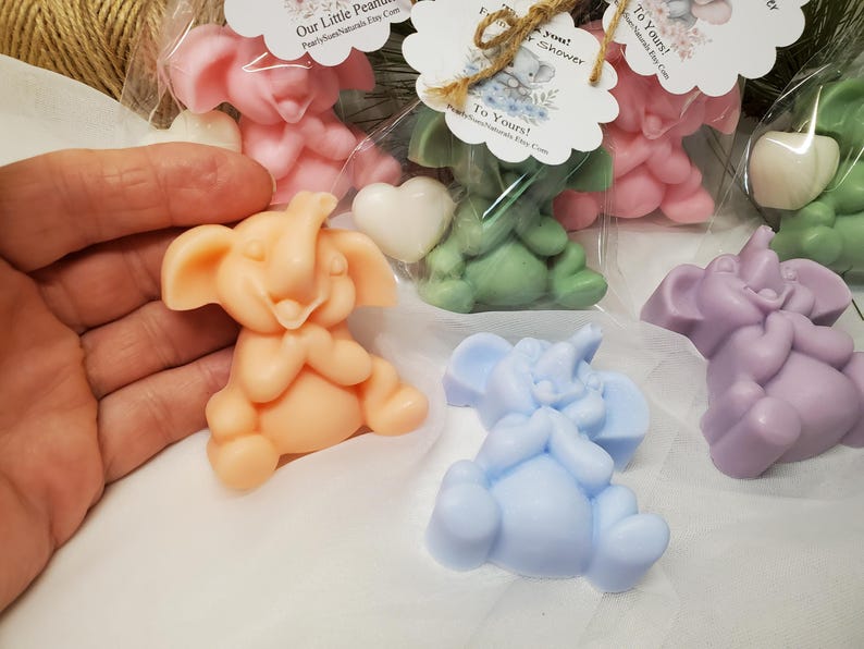 May include: A variety of colorful elephant-shaped soaps in pink, green, blue, and purple. Each soap is a sitting elephant with a smiling face. Some are packaged with a heart-shaped soap and a tag.