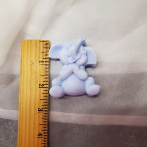 May include: A light blue elephant figurine, approximately 2 inches tall, in a seated position with its trunk raised. The elephant is made of a smooth material and has a smiling expression. A wooden ruler is next to the elephant for size comparison.