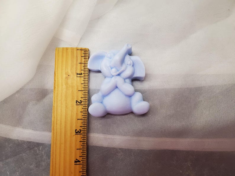 May include: A light blue elephant figurine, approximately 2 inches tall, in a seated position with its trunk raised. The elephant is made of a smooth material and has a smiling expression. A wooden ruler is next to the elephant for size comparison.