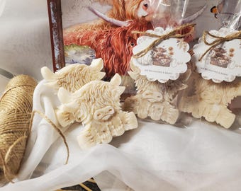 May include: Cream-colored Highland cow-shaped soaps. Two soaps are packaged with tags that read "Holy Cow We're Having a Baby!" A spool of twine and a watercolor painting of a Highland cow are in the background.