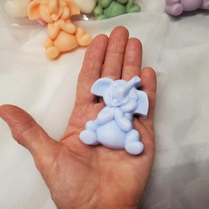 May include: A light blue elephant-shaped soap figurine. The soap is in the shape of a sitting elephant with its trunk raised. Other elephant soaps in peach, green, and purple are visible in the background.