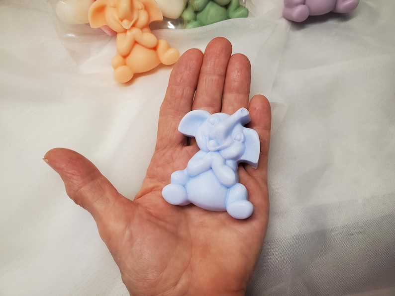 May include: A light blue elephant-shaped soap figurine. The soap is in the shape of a sitting elephant with its trunk raised. Other elephant soaps in peach, green, and purple are visible in the background.
